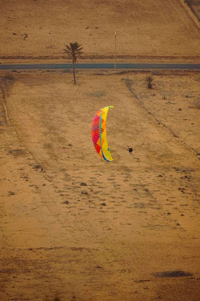 A vibrant paraglider soaring over a vast desert terrain, aerial view.